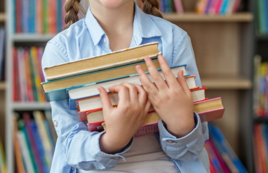 Dziewczynka w niebieskiej koszuli trzyma stos książek, uśmiechając się w bibliotece.