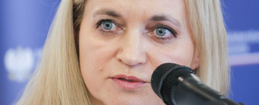 Portret kobiety z jasnymi włosami, przy mikrofonie, w tle niebieska ściana.