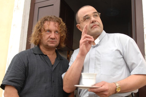 Dwaj mężczyźni stoją przy drzwiach. Jeden trzyma filiżankę, drugi wskazuje palcem.