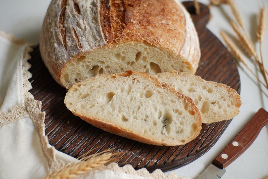 Chleb na drewnianej desce, kilka kromek obok bochenka, w tle zboża i nóż.
