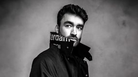 HI-LO w czarnej kurtce z wysokim kołnierzem, patrzy z lekkim uśmiechem. Czarno-biała fotografia.