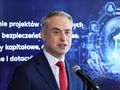 Mężczyzna w garniturze z czerwonym krawatem, przemawia do mikrofonu. W tle tekst o projektach.