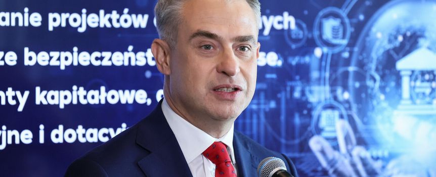 Mężczyzna w garniturze z czerwonym krawatem, przemawia do mikrofonu. W tle tekst o projektach.