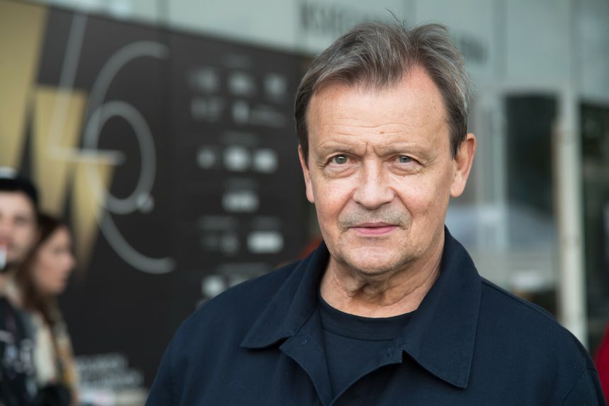 Mężczyzna z krótkimi, siwymi włosami w granatowej kurtce, uśmiechający się do kamery.