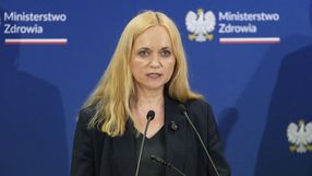 Kobieta z długimi blond włosami przemawia przy mikrofonach, w tle logo Ministerstwa Zdrowia.