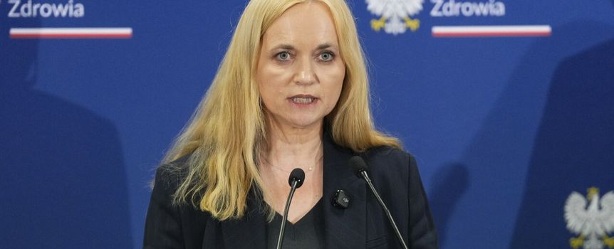 Kobieta z długimi blond włosami przemawia przy mikrofonach, w tle logo Ministerstwa Zdrowia.