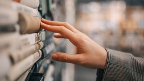 Zdjęcie przedstawia dłoń dotykającą książek na półce w bibliotece.
