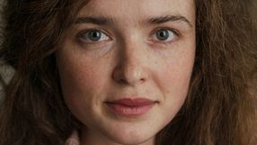 Portret młodej kobiety z brązowymi włosami i zielonymi oczami, uśmiechającej się delikatnie.