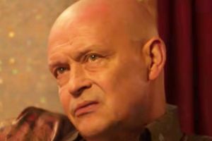 Mężczyzna z krótkimi włosami, poważnym wyrazem twarzy, na tle kolorowej ścianki.