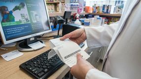 Osoba w białym fartuchu trzyma paragon przy komputerze w aptece. W tle produkty apteczne.