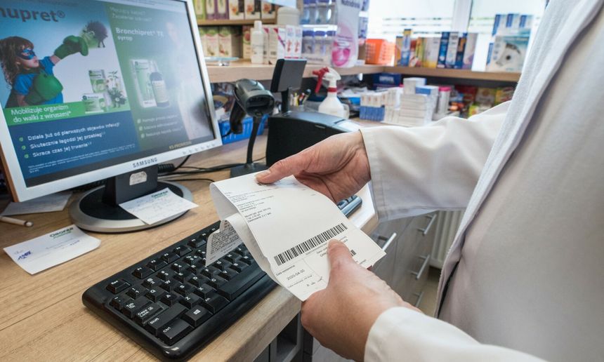 Osoba w białym fartuchu trzyma paragon przy komputerze w aptece. W tle produkty apteczne.