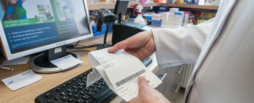Osoba w białym fartuchu trzyma paragon przy komputerze w aptece. W tle produkty apteczne.
