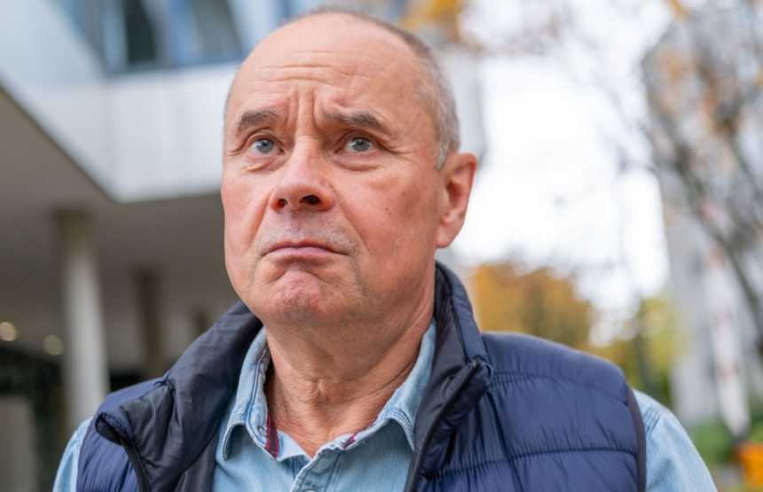 Mężczyzna z poważnym wyrazem twarzy, ubrany w granatową kamizelkę. Tło rozmyte.