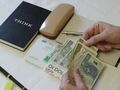 Na stole leżą pieniądze, notatnik, stempel i długopis; ręka trzyma banknoty.