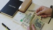 Na stole leżą pieniądze, notatnik, stempel i długopis; ręka trzyma banknoty.