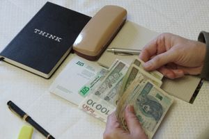 Na stole leżą pieniądze, notatnik, stempel i długopis; ręka trzyma banknoty.