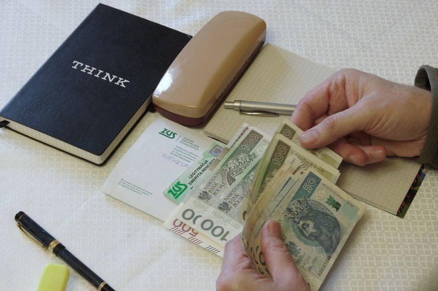 Na stole leżą pieniądze, notatnik, stempel i długopis; ręka trzyma banknoty.