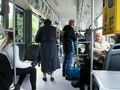 Wnętrze autobusu, pasażerowie siedzą i stoją, jedna osoba w płaszczu wychodzi.