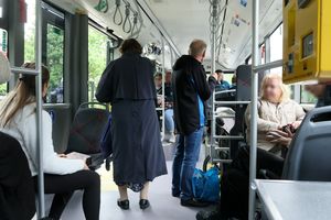 Wnętrze autobusu, pasażerowie siedzą i stoją, jedna osoba w płaszczu wychodzi.