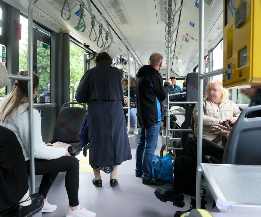 Wnętrze autobusu, pasażerowie siedzą i stoją, jedna osoba w płaszczu wychodzi.