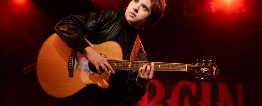 Młody mężczyzna w czarnym ubraniu gra na gitarze na scenie z czerwonym oświetleniem.