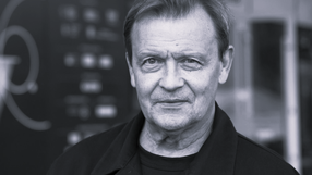 Zdjęcie przedstawia mężczyznę w średnim wieku z uśmiechem, w czarnym ubraniu, na tle rozmytych elementów.