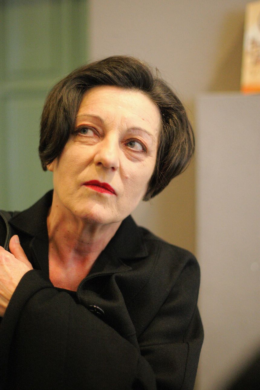 Kobieta w czarnym płaszczu, trzymająca się za ramię, z wyrazem niepokoju na twarzy.