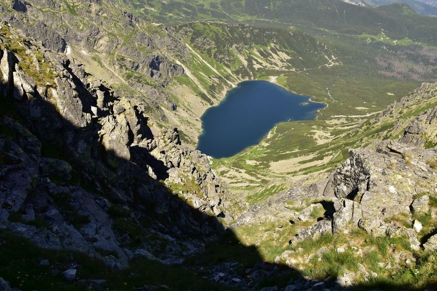 Widok z góry na dolinę z jeziorem, otoczoną stromymi skałami i zielonymi łąkami.
