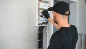 Mężczyzna w czapce naprawia rozdzielnicę elektryczną na szarej ścianie.