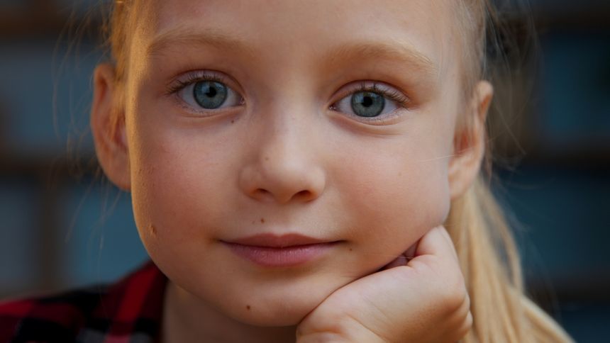 Portret dziewczynki z niebieskimi oczami, opierającej głowę na ręce. Uśmiech w tle.
