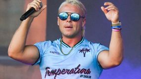 Skolim z blond włosami w okularach przeciwsłonecznych, trzyma mikrofon i uśmiecha się.