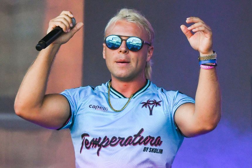 Skolim z blond włosami w okularach przeciwsłonecznych, trzyma mikrofon i uśmiecha się.