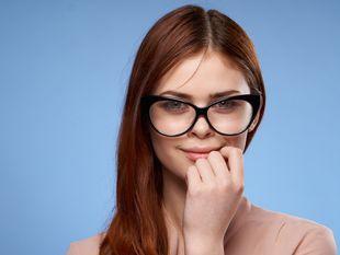 Młoda kobieta z długimi, rudymi włosami i dużymi okularami, z uśmiechem i lekko podniesioną dłonią.