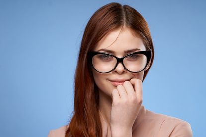 Młoda kobieta z długimi, rudymi włosami i dużymi okularami, z uśmiechem i lekko podniesioną dłonią.