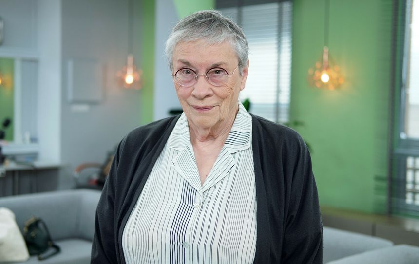 Starsza kobieta z krótkimi, szarymi włosami, w okularach, uśmiecha się w studiu telewizyjnym.