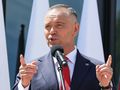 Mężczyzna w garniturze z mikrofonem, przemawia, w tle biało-czerwone flagi.