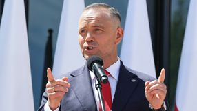 Mężczyzna w garniturze z mikrofonem, przemawia, w tle biało-czerwone flagi.