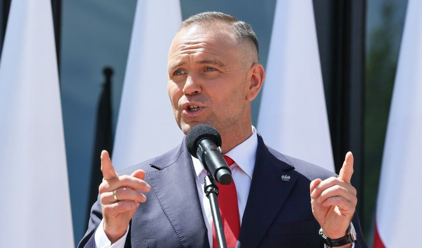 Mężczyzna w garniturze z mikrofonem, przemawia, w tle biało-czerwone flagi.