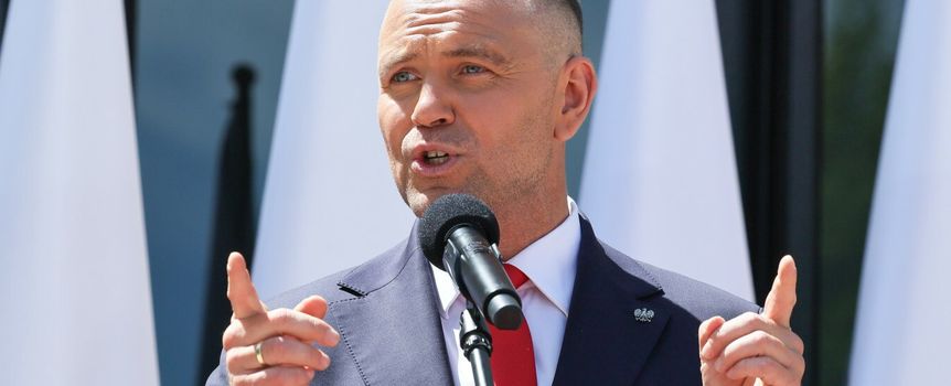 Mężczyzna w garniturze z mikrofonem, przemawia, w tle biało-czerwone flagi.