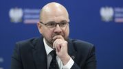 Mężczyzna w okularach w garniturze, z zamyśloną miną, siedzi przy stole. Tło w kolorze niebieskim.