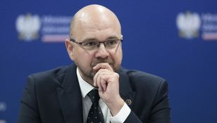 Mężczyzna w okularach w garniturze, z zamyśloną miną, siedzi przy stole. Tło w kolorze niebieskim.