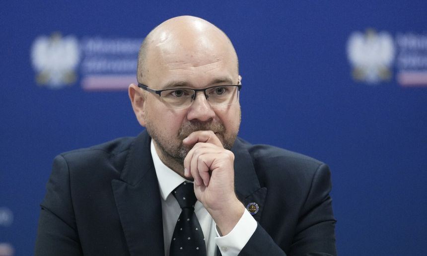 Mężczyzna w okularach w garniturze, z zamyśloną miną, siedzi przy stole. Tło w kolorze niebieskim.