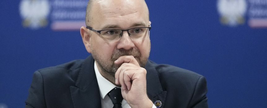 Mężczyzna w okularach w garniturze, z zamyśloną miną, siedzi przy stole. Tło w kolorze niebieskim.
