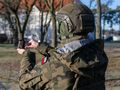 Żołnierz w mundurze kamuflażowym trzyma mały ekran w dłoni. W tle drzewa i budynki.