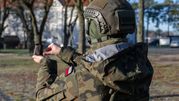 Żołnierz w mundurze kamuflażowym trzyma mały ekran w dłoni. W tle drzewa i budynki.