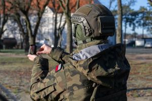 Żołnierz w mundurze kamuflażowym trzyma mały ekran w dłoni. W tle drzewa i budynki.