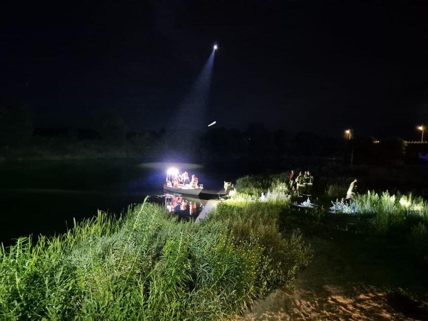Na zdjęciu łódź na spokojnej wodzie w nocy, światła oraz osoby na brzegu.