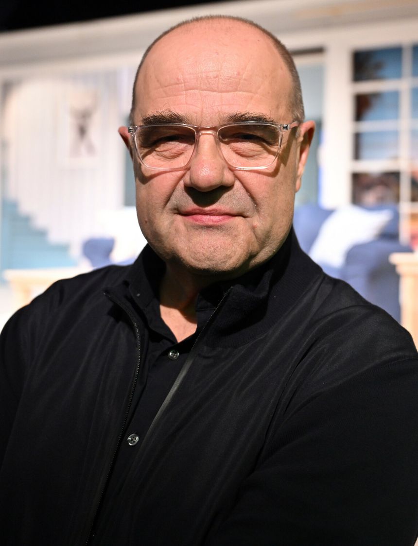 Mężczyzna z łysą głową, w okularach, stoi na tle jasnego pomieszczenia. Uśmiecha się.