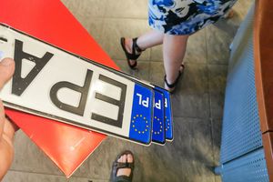 Na zdjęciu ręka trzyma trzy tablice rejestracyjne oraz widoczna część nogi innej osoby.