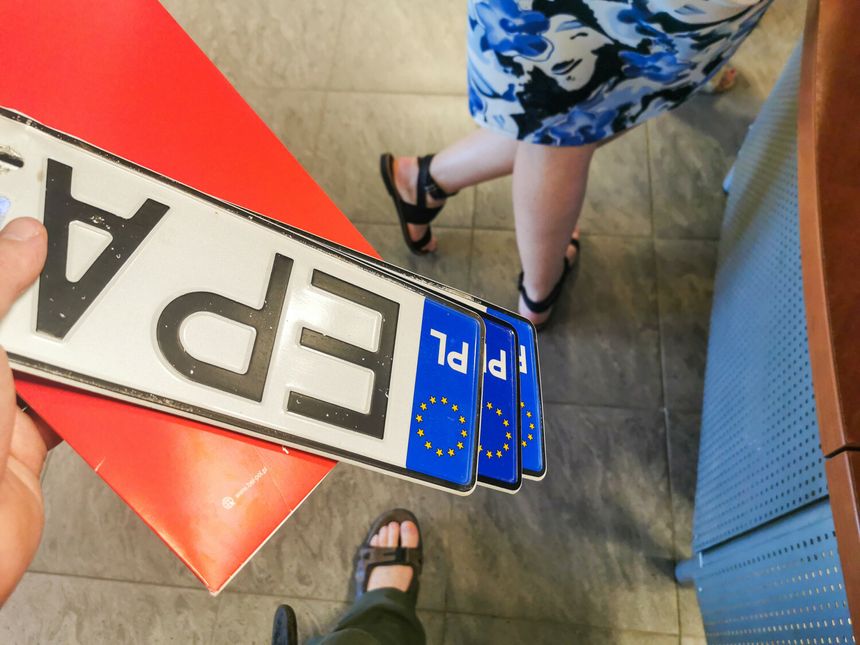 Na zdjęciu ręka trzyma trzy tablice rejestracyjne oraz widoczna część nogi innej osoby.